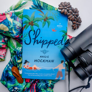 Shipped by Angie Hockman lays on top of a tropical printed scarf with a wooden flower hair clip above it and binoculars to the left of the book.